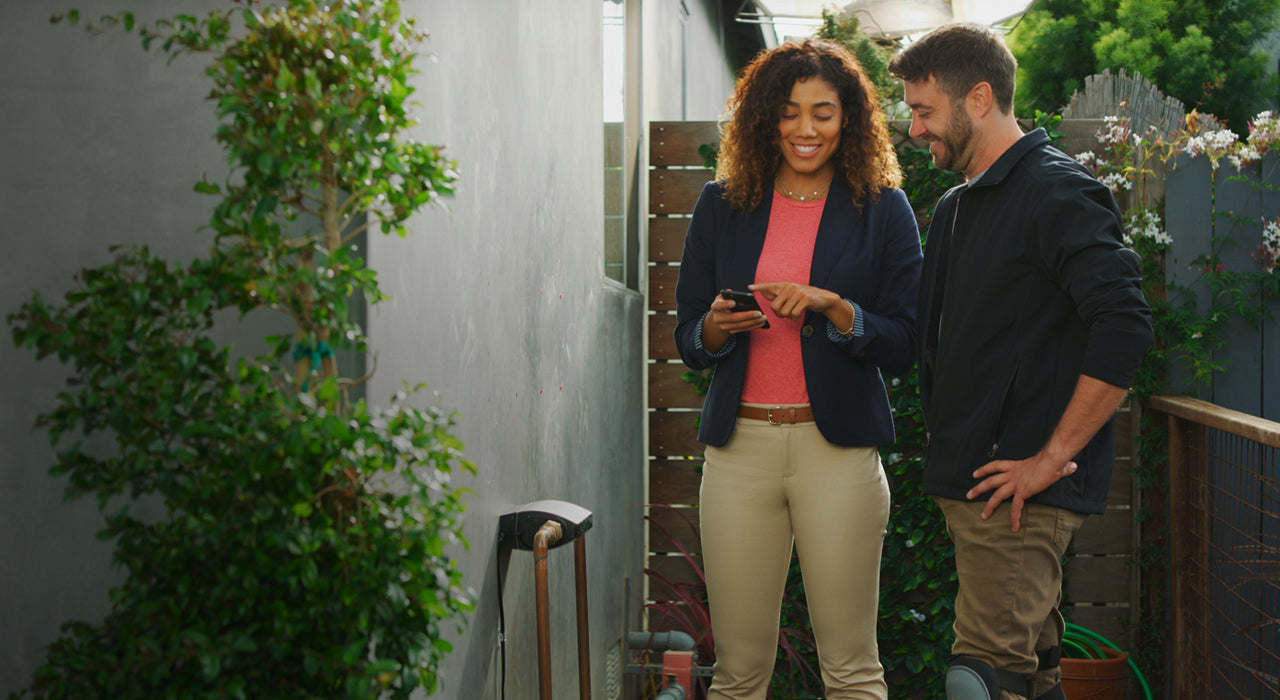 A woman smiling and pointing to a smart phone next to a man installing a Phyn device.